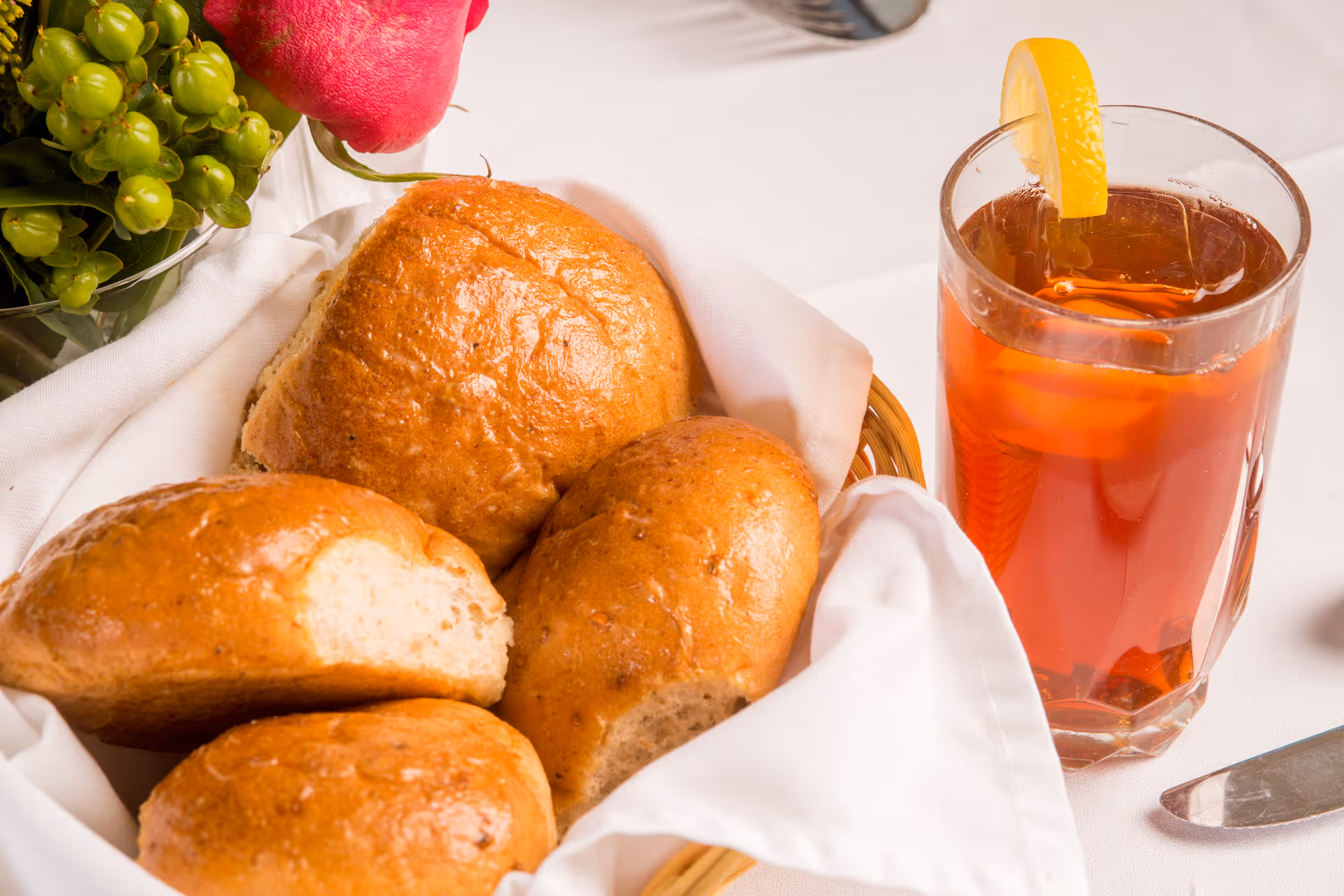 Basket of bread rolls on a napkin beside a glass of iced tea garnished with a lemon slice on a white tablecloth.
