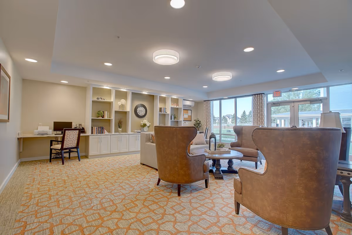 Bright communal lounge with leather armchairs around a coffee table, built-in shelving, a desk area, and large windows.