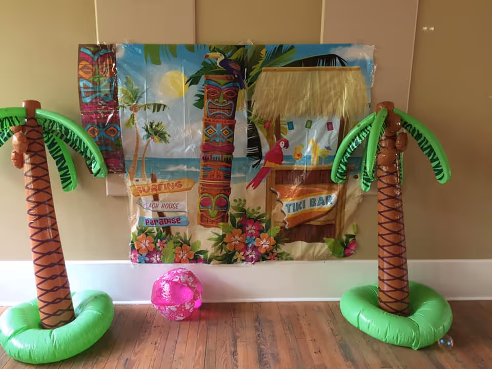 Two inflatable palm trees and a pink inflatable beach ball placed on a wooden floor in front of a wall decorated with a colorful tropical-themed backdrop featuring tiki totems, palm trees, flowers, a parrot, and signs reading 'Surfing', 'Beach House', 'Paradise', and 'Tiki Bar'.
