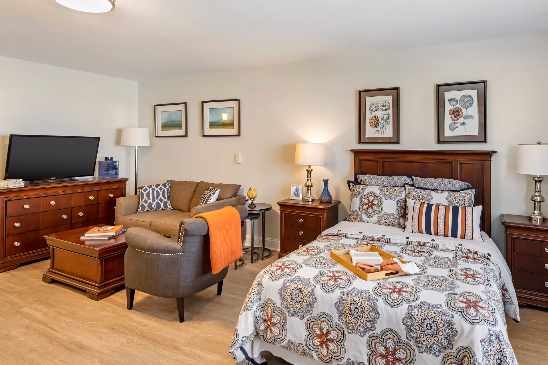 A well-appointed studio suite with a bed and nightstands, a seating area with a sofa and armchair, a TV on a dresser, and framed artwork on the walls.