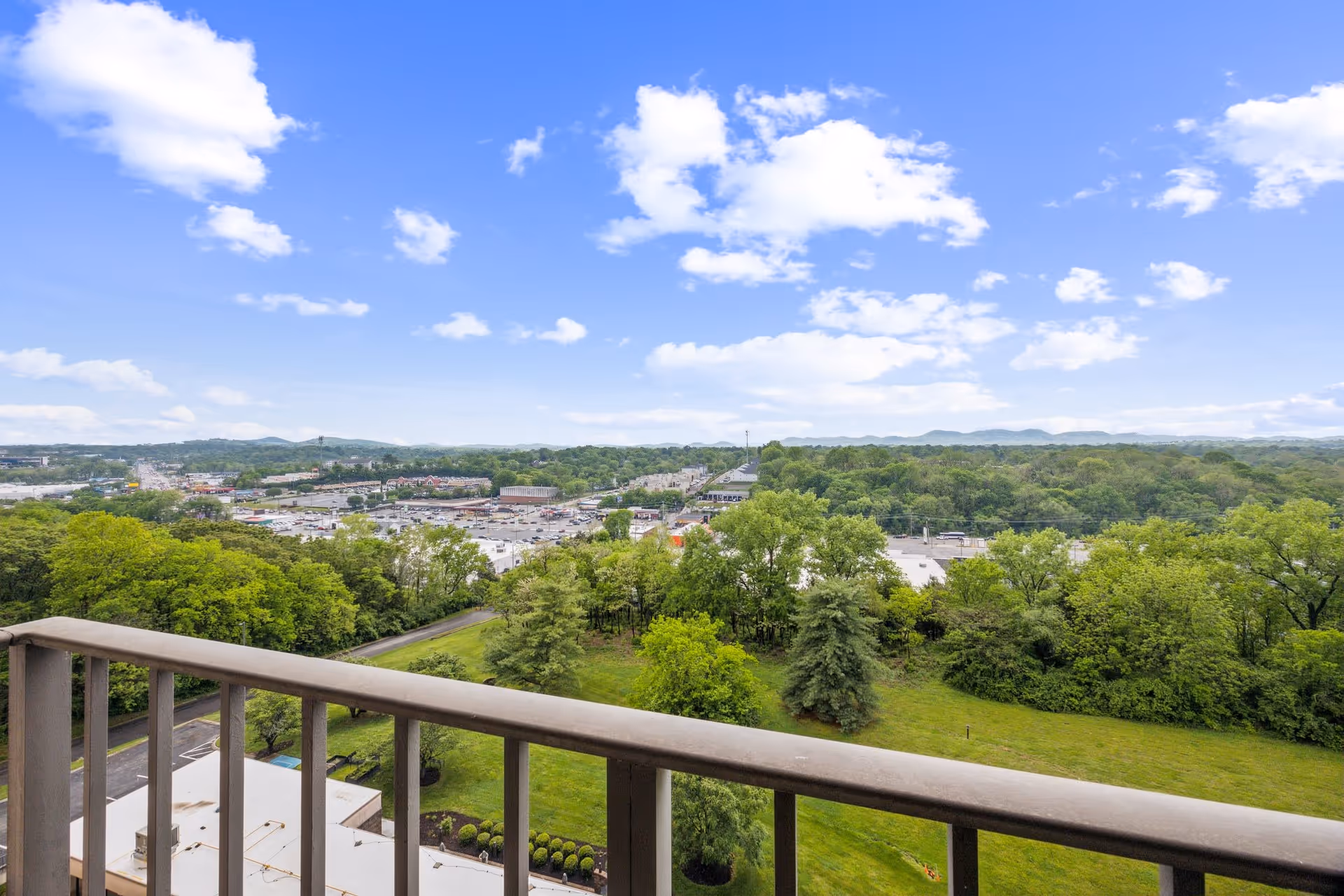 View from a balcony overlooking a green landscape with trees, a grassy area, and a distant view of a town and hills under a partly cloudy blue sky.