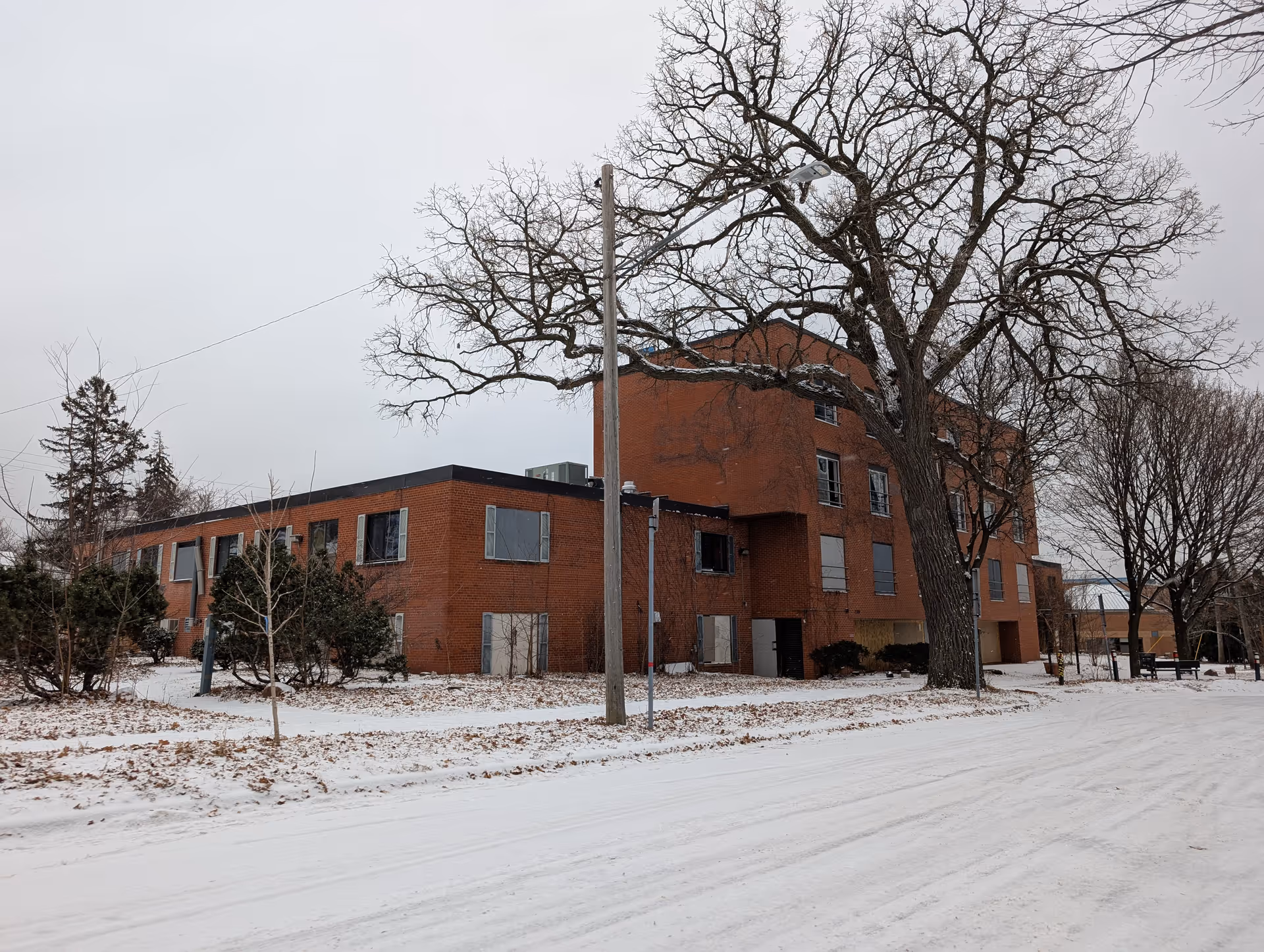 Exterior view of a multi-story brick building in winter with snow covering the ground and leafless trees around. The building has several windows, some boarded up, and a large tree in the foreground.