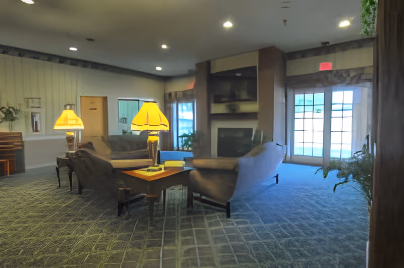 A cozy living room area with carpeted flooring, featuring a wooden coffee table with a lamp on it, surrounded by upholstered armchairs and sofas. There is a fireplace with a mounted TV above it, large windows and glass doors letting in natural light, and some green plants near the entrance.