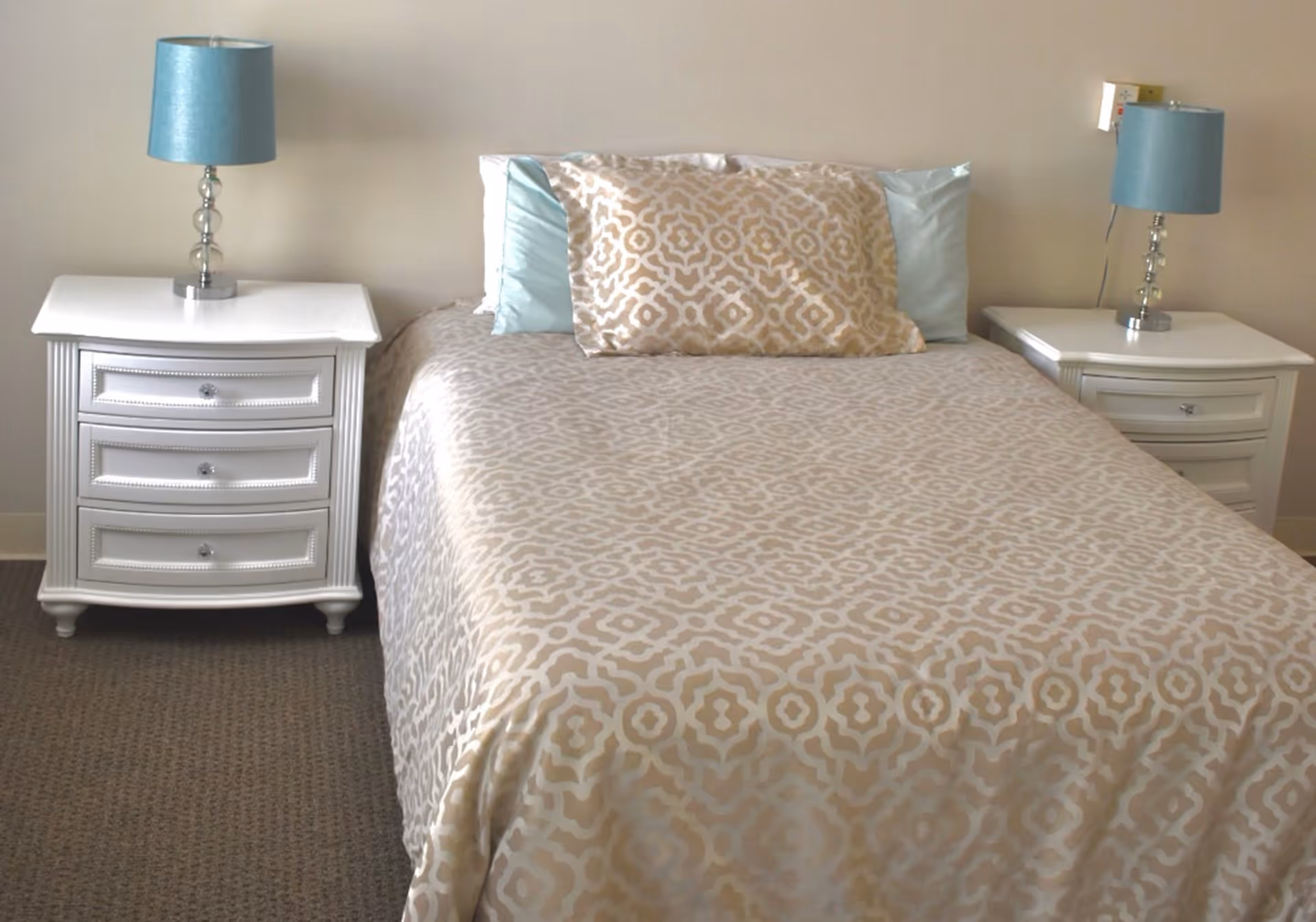 A neatly made bed with a beige and white patterned bedspread and matching pillow, flanked by two white nightstands each with a blue lampshade on top, set against a plain light-colored wall.