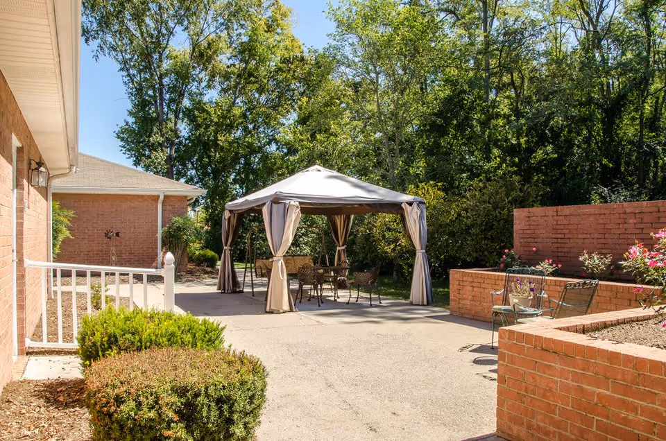Outdoor patio area at Azalea Court Senior Living featuring a gazebo with curtains, metal chairs and tables, surrounded by brick planters with flowers and greenery, and trees in the background under a clear sky.