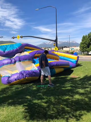 A deflated colorful inflatable slide on a grassy lawn with a person standing next to it and mountains under a blue sky in the background.