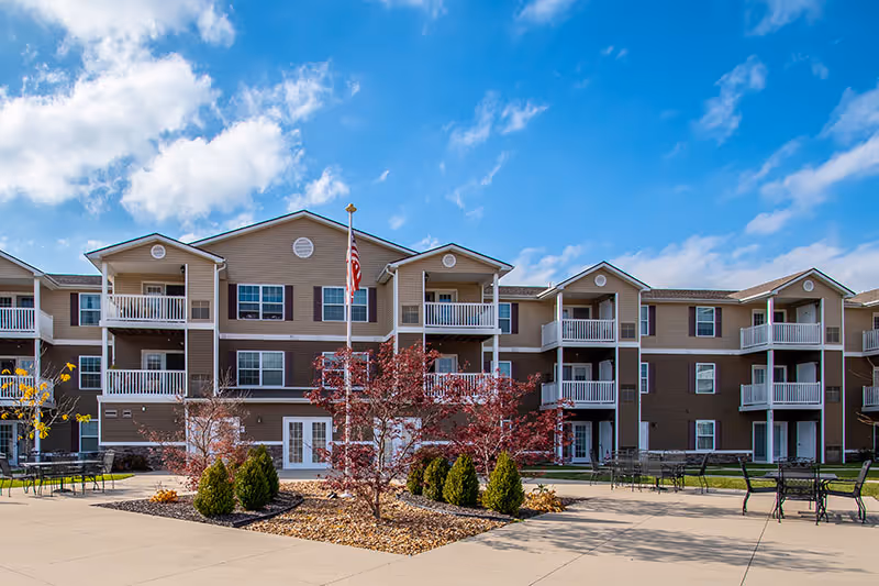 Three-story senior living building with balconies overlooking a landscaped courtyard and an American flag under a blue sky.