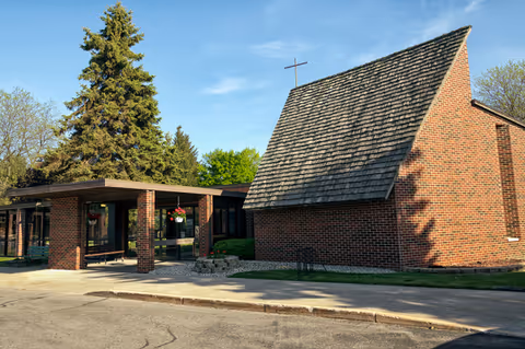 Brick retirement community building with covered entrance, cross on roof, and trees nearby under blue sky.