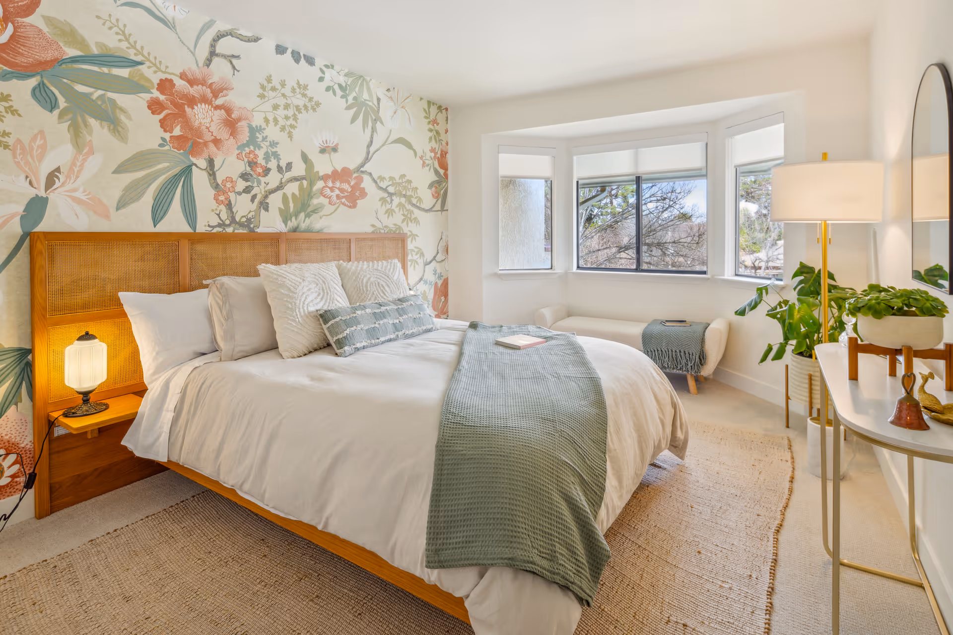 A bright and cozy bedroom featuring a large bed with white bedding and decorative pillows, a green throw blanket, a wooden headboard, and a floral accent wall behind the bed. There is a window with a view of trees, a small white bench with a green throw, a white console table with plants and decorative items, and a tall floor lamp providing warm light.