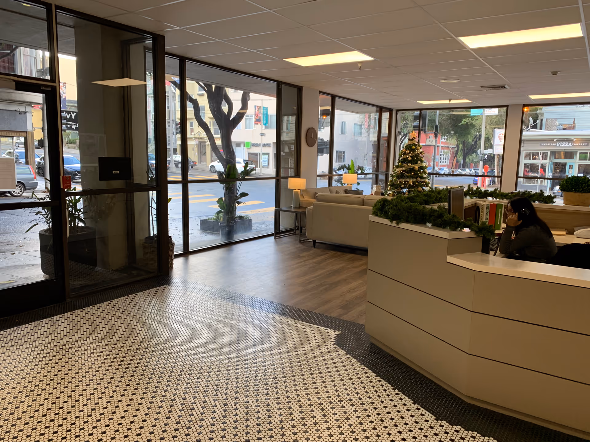 Interior view of a facility lobby with large windows showing the street outside. There is a reception desk decorated with green garlands and a small Christmas tree. A person is sitting at the desk on the phone. Comfortable seating with sofas and lamps is visible in the background.