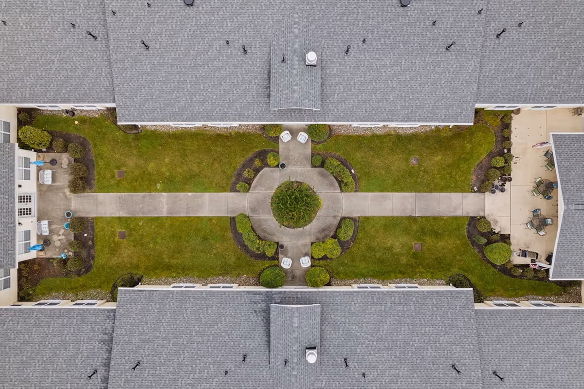 Aerial view of a senior living facility courtyard with a central circular walkway surrounded by green grass and shrubs. The courtyard is bordered by buildings with gray shingled roofs, and there are outdoor seating areas with chairs and tables on both ends of the courtyard.