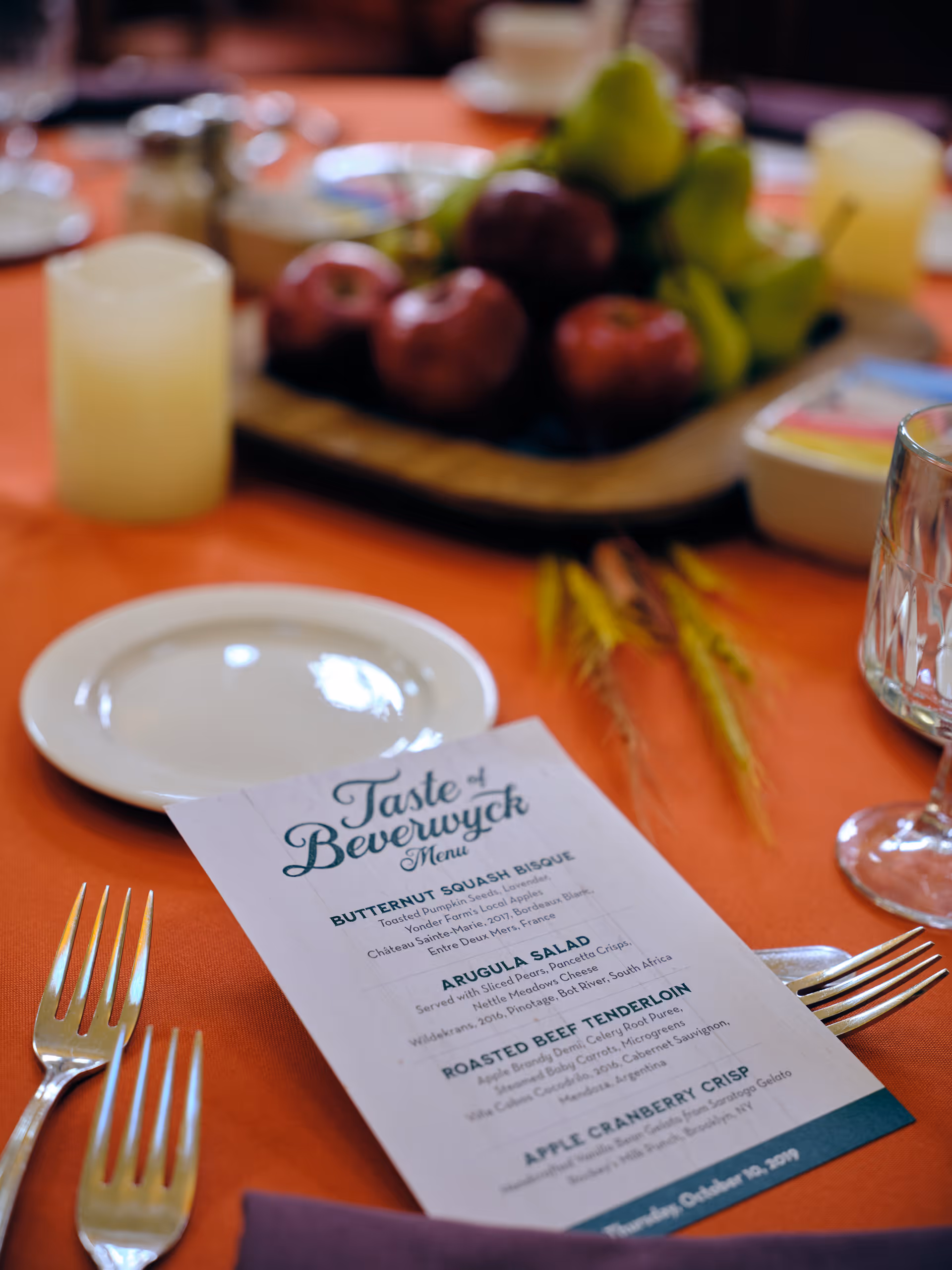 A close-up view of a dining table set with an orange tablecloth, a white plate, two forks, a glass, and a menu titled 'Taste of Beverwyck'. In the background, there is a wooden tray with apples and pears, along with other dining items slightly out of focus.