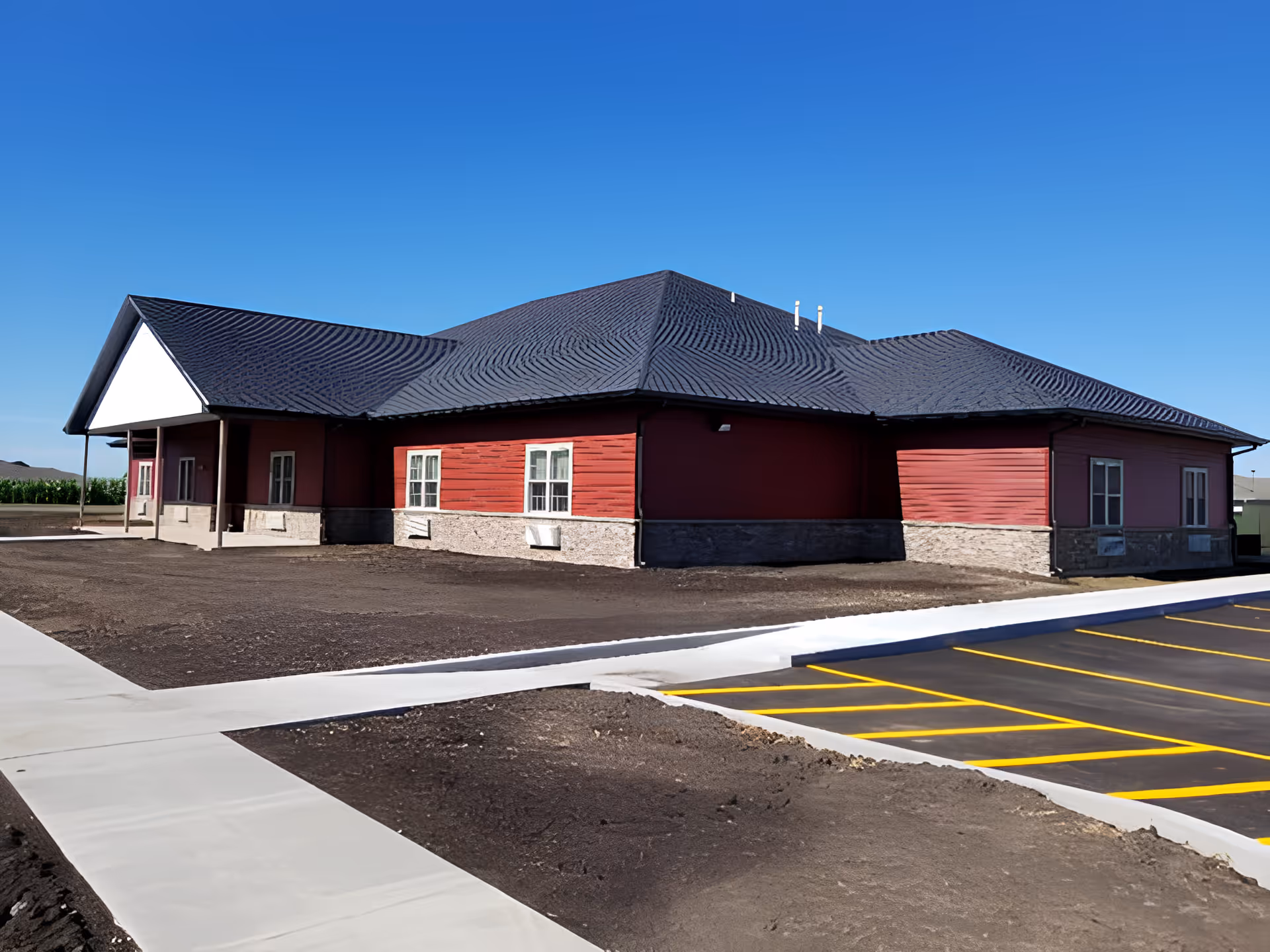 Single-story red assisted living building with a dark roof, adjacent paved parking area and sidewalks under a clear blue sky.