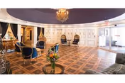 A spacious, elegant common area with a polished wooden parquet floor, several blue upholstered chairs with wooden frames arranged around small tables, a chandelier hanging from a domed ceiling, and large windows allowing natural light to fill the room.