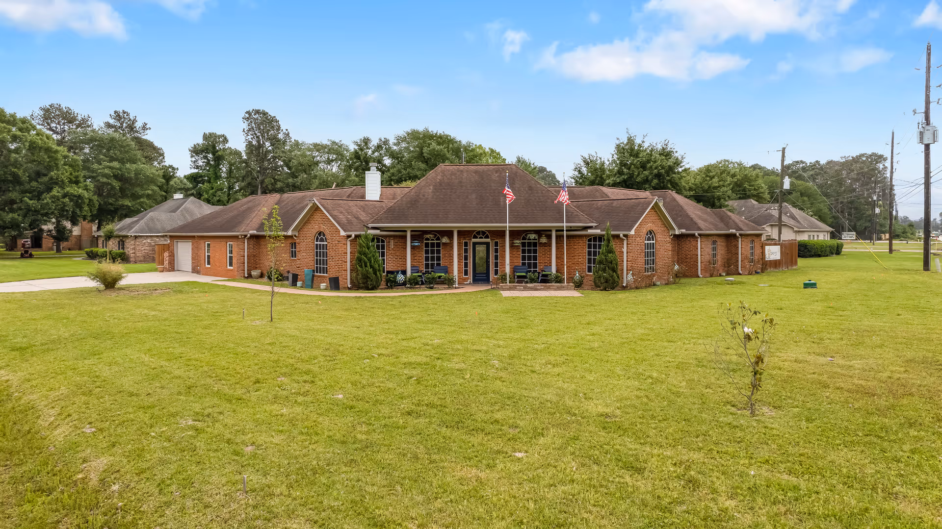 Front exterior view of a single-story brick building with a brown roof, two American flags on poles in front, surrounded by a large green lawn and trees in the background under a blue sky with some clouds.