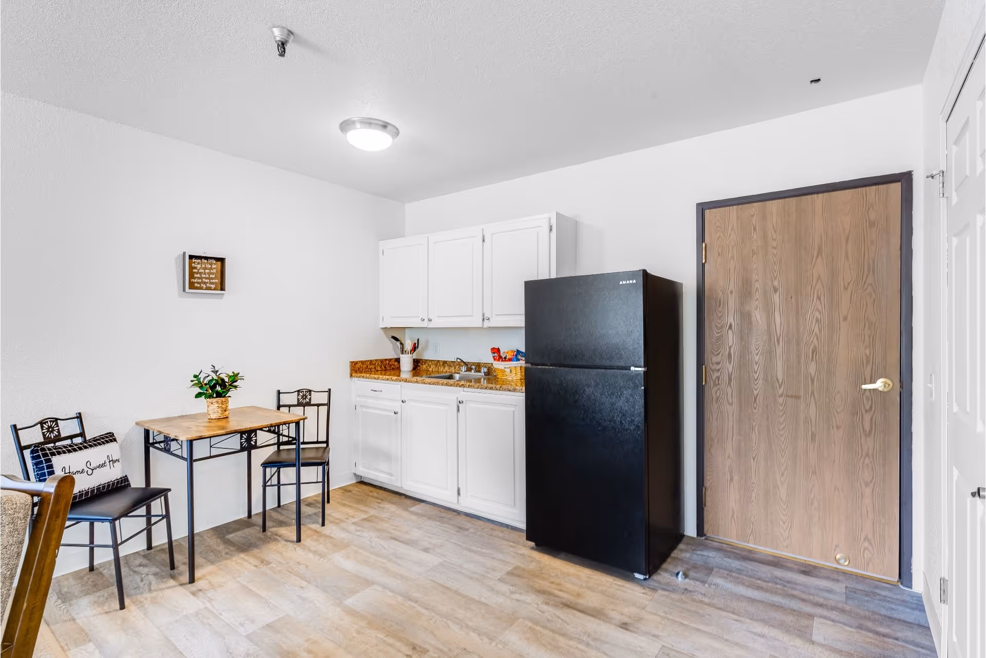 A small kitchen area with white cabinets, a black refrigerator, and a granite countertop with a sink. To the left, there is a small wooden table with two metal chairs, one with a 'Home Sweet Home' pillow. The floor is light wood, and there is a wooden door on the right side of the image.