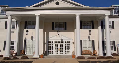 Front exterior view of a large building with white columns, multiple windows with black shutters, and a central entrance with glass double doors. There are benches and potted plants near the entrance, and the sky is clear and blue.