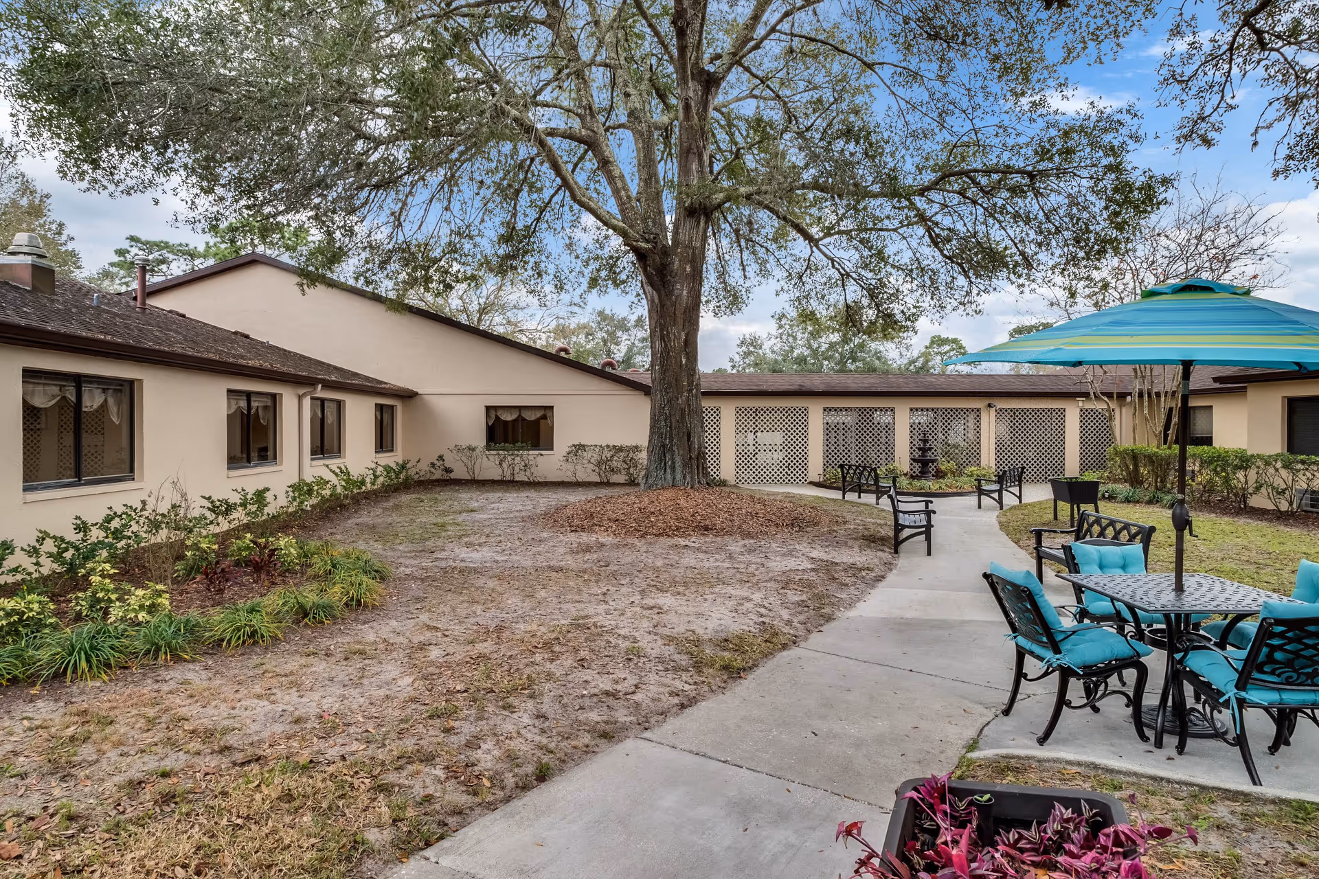 Courtyard of a senior living facility with a large oak tree, a paved walkway, turquoise-padded patio tables and chairs, and surrounding single-story beige buildings.