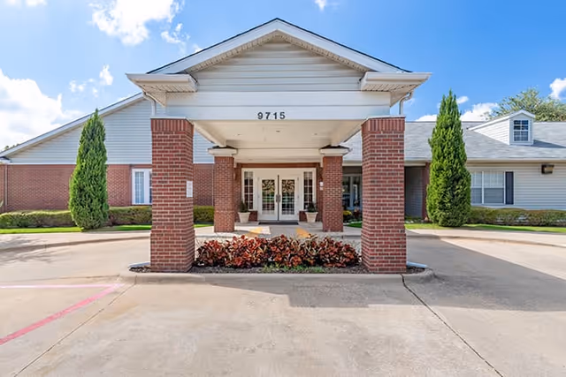 Front exterior view of a single-story building with a covered entrance supported by four brick columns. The building has white siding and brick walls, with the number 9715 displayed above the entrance. There are two tall evergreen shrubs on either side of the entrance and a small landscaped area with red and green plants in front of the columns. The sky is blue with some clouds.