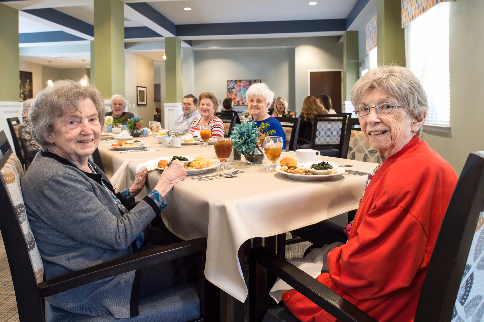A group of elderly people sitting around a dining table in a well-lit room, enjoying a meal together. The table is covered with a beige tablecloth and set with plates of food, glasses of iced tea, and a small floral centerpiece. The room has green walls and large windows with patterned curtains.