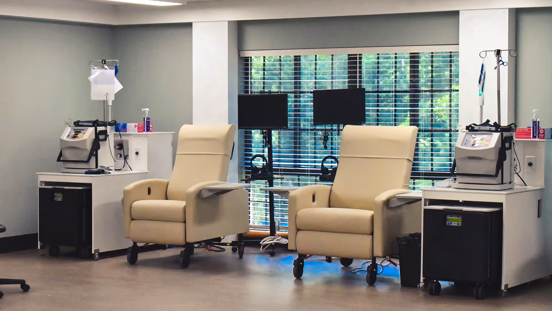 A medical treatment room with two beige reclining chairs on wheels, each positioned next to a medical cart with equipment and IV stands. Behind the chairs are two monitors mounted on stands in front of a large window with horizontal blinds, allowing natural light to enter.