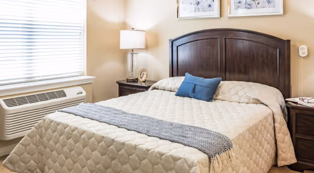 A neatly made bed with a beige quilted bedspread and a blue accent pillow in the center. The bed has a dark wooden headboard. On either side of the bed are matching dark wooden nightstands, one with a lamp and a small framed photo, and the other with a folded towel. A window with white blinds is visible on the left side of the room, above a wall-mounted air conditioning unit.