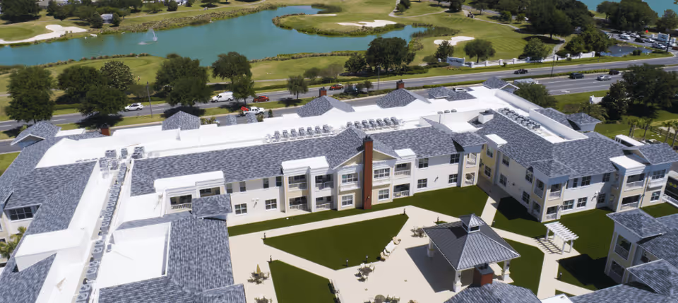 Aerial view of Watercrest Buena Vista senior living facility showing a large building with gray shingled roofs surrounding a green courtyard with pathways, seating areas, and a gazebo. Beyond the building, there is a road with cars and a golf course with a pond and fountain in the background.