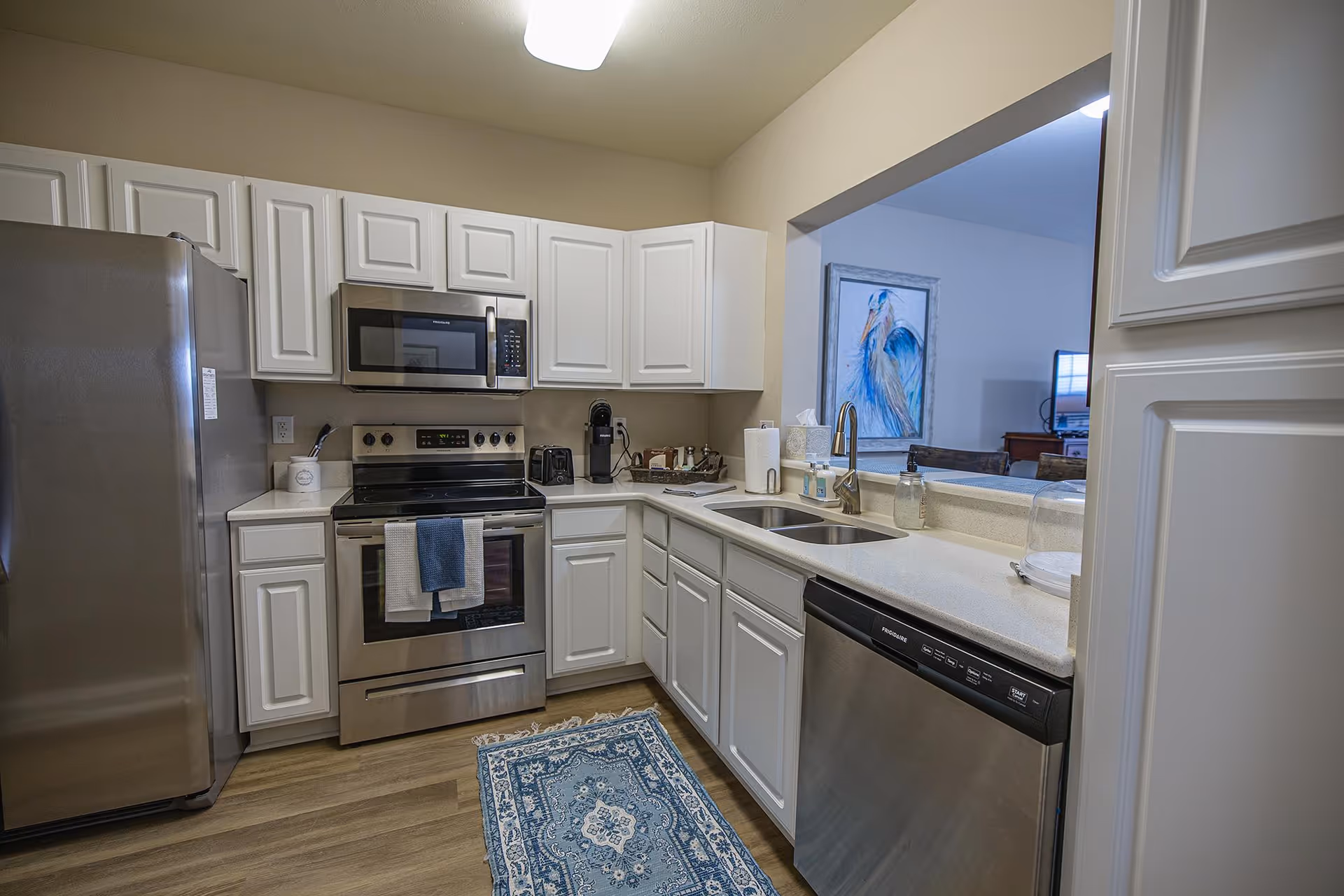 Modern kitchen with white cabinets, stainless steel refrigerator, oven, microwave, and dishwasher. There is a blue patterned rug on the wooden floor and a window opening to an adjacent room with a painting of a blue bird on the wall.