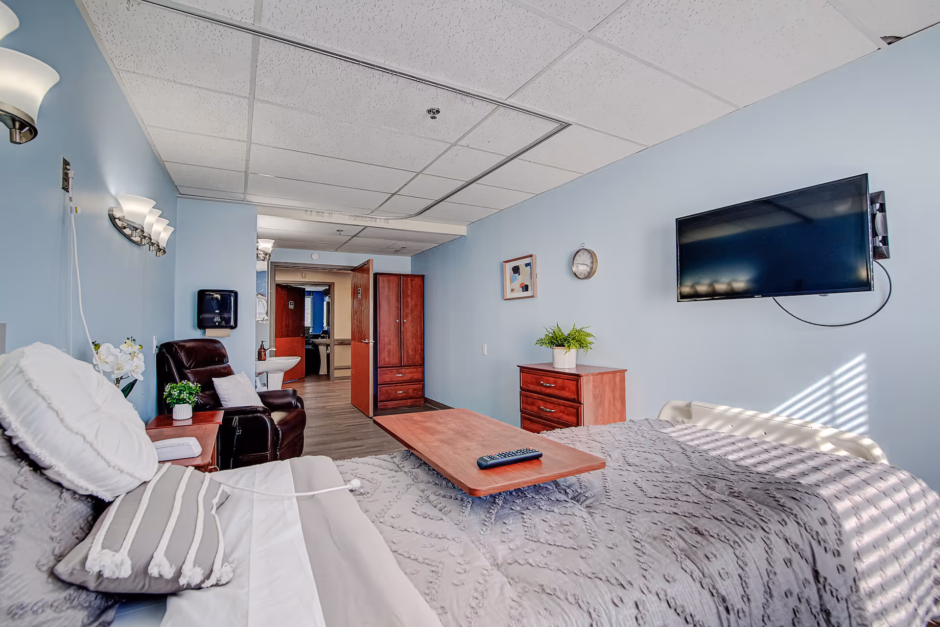 A senior living facility bedroom with a bed covered in a gray patterned blanket and pillows, a wooden table with a remote control on it, a leather recliner chair with a pillow, a small wooden nightstand with a plant and flowers, a wall-mounted TV, a wooden dresser with a plant on top, a clock and framed artwork on the light blue walls, and an open doorway leading to a bathroom area.