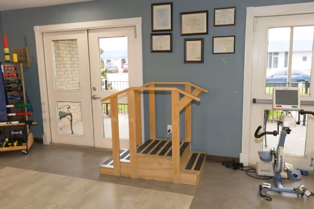 Rehabilitation room with wooden parallel bars and steps, an exercise bike, weight rack, and framed certificates on the wall.