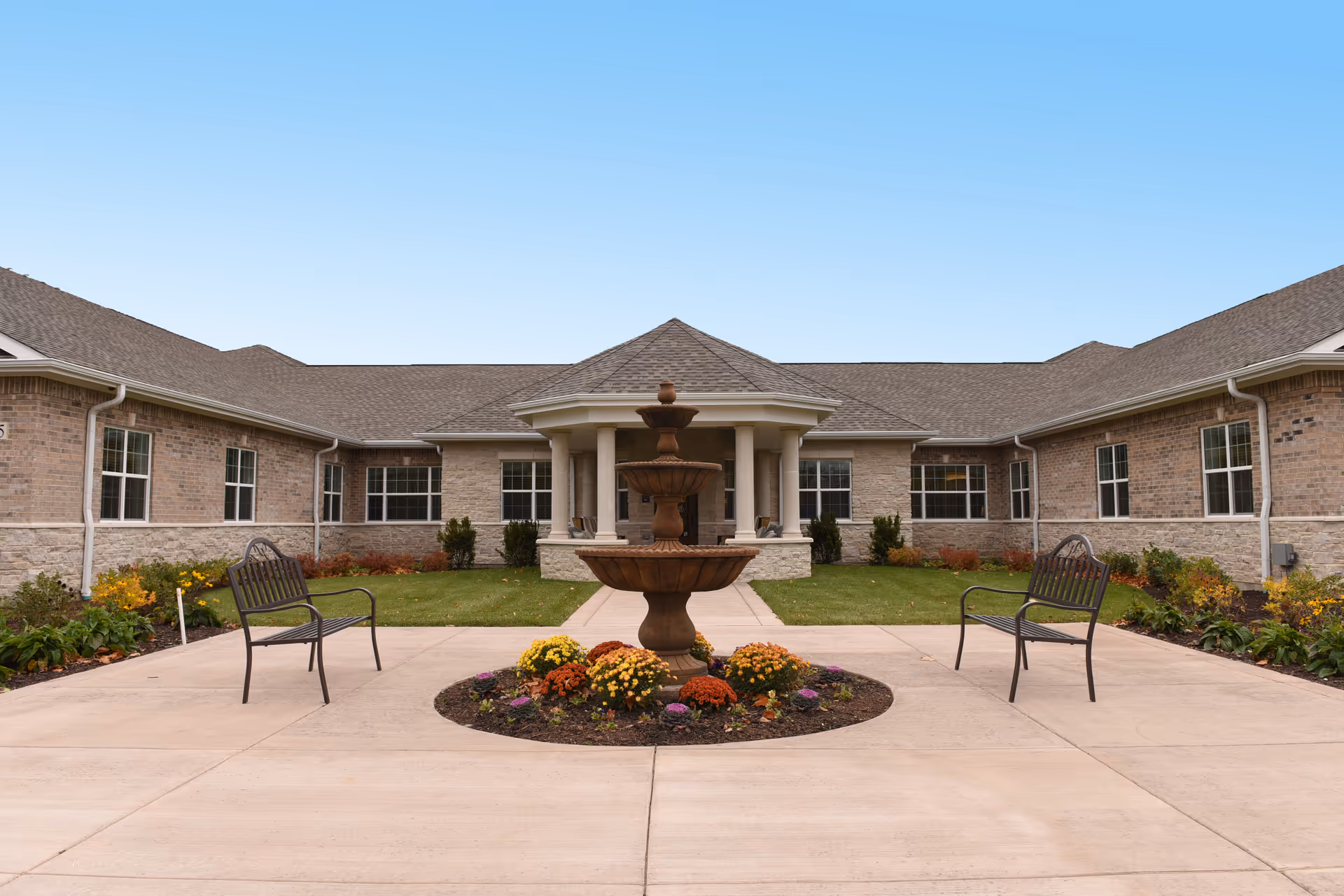 Front courtyard of a single-story brick memory care building featuring a central tiered fountain, flowerbeds, and benches.