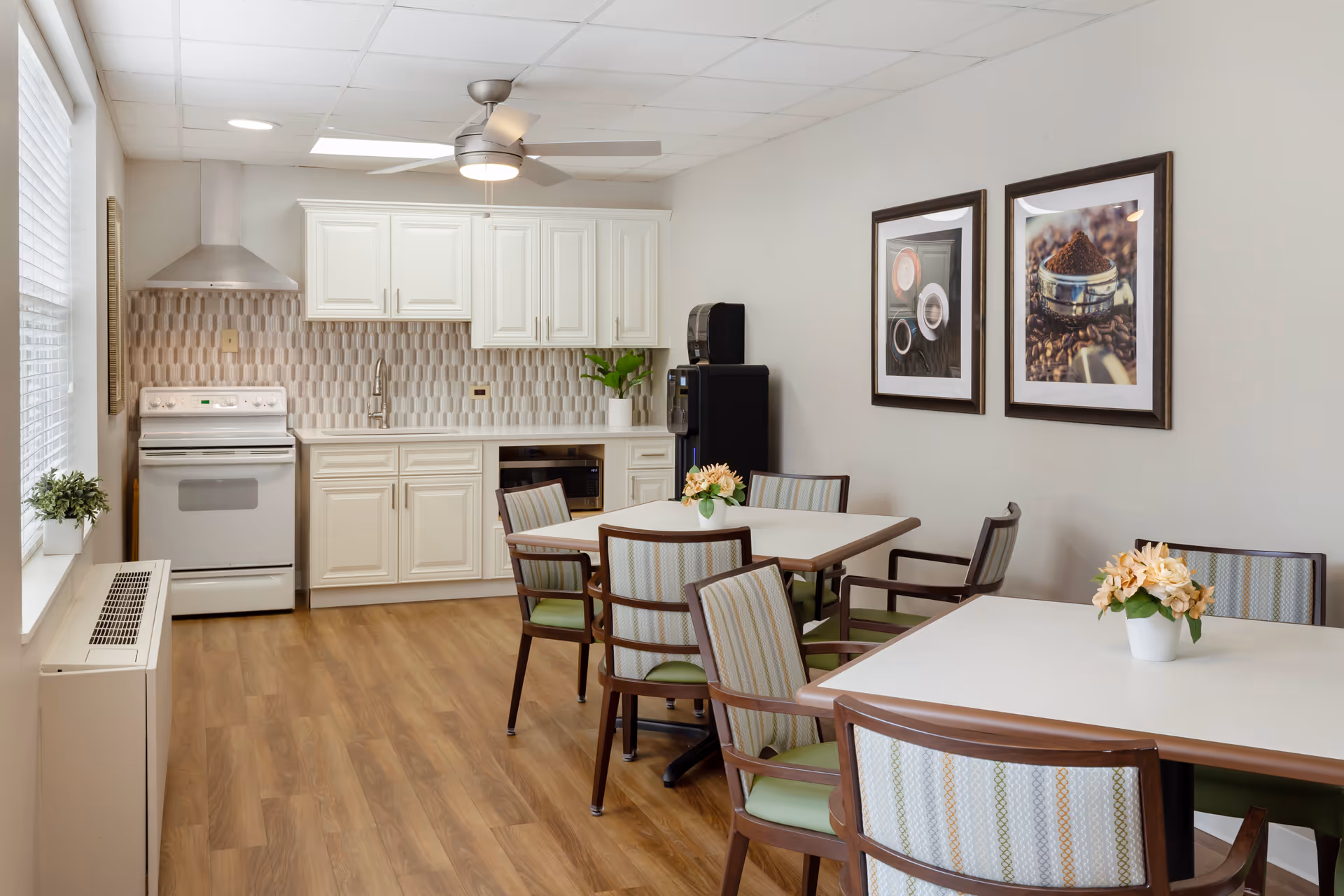 A bright kitchen and dining area with white cabinets, a stove, a microwave, and a water dispenser. There are two tables with chairs around them, each table decorated with a small flower arrangement. The walls are adorned with two framed pictures of coffee-related items. The floor is wooden, and a ceiling fan with a light is mounted above.