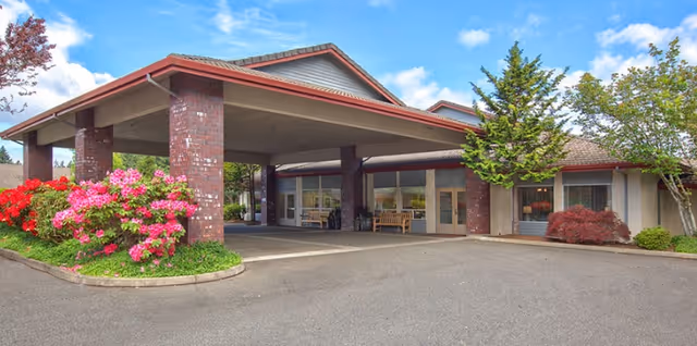 Exterior view of Alpine Way Continuing Care Community featuring a covered entrance supported by brick pillars, surrounded by landscaped bushes and flowering plants under a partly cloudy blue sky.