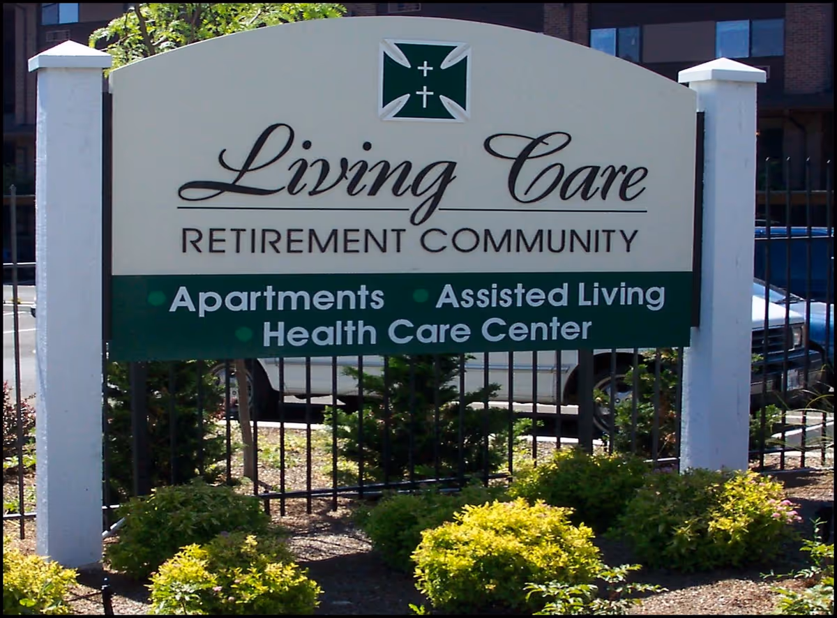 Outdoor sign for Living Care Retirement Community featuring services such as Apartments, Assisted Living, and Health Care Center, surrounded by bushes and a black metal fence.