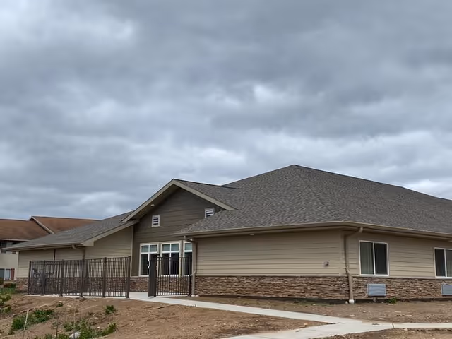Exterior view of a single-story building with beige siding and stone accents under a cloudy sky. The building has a gray shingled roof and several windows. There is a black metal fence and a concrete walkway leading to the entrance.