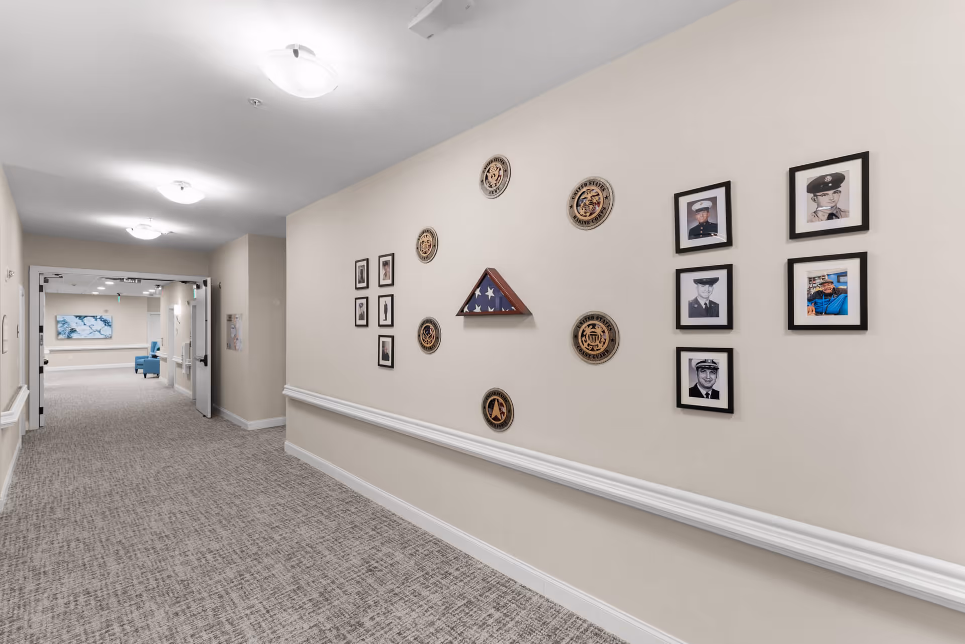 A well-lit hallway in a senior living facility with beige walls and carpeted floor. The right wall features framed photographs of military personnel and circular plaques with military insignias, along with a folded American flag in a triangular display case. The hallway leads to an open area with blue chairs and a large artwork on the far wall.