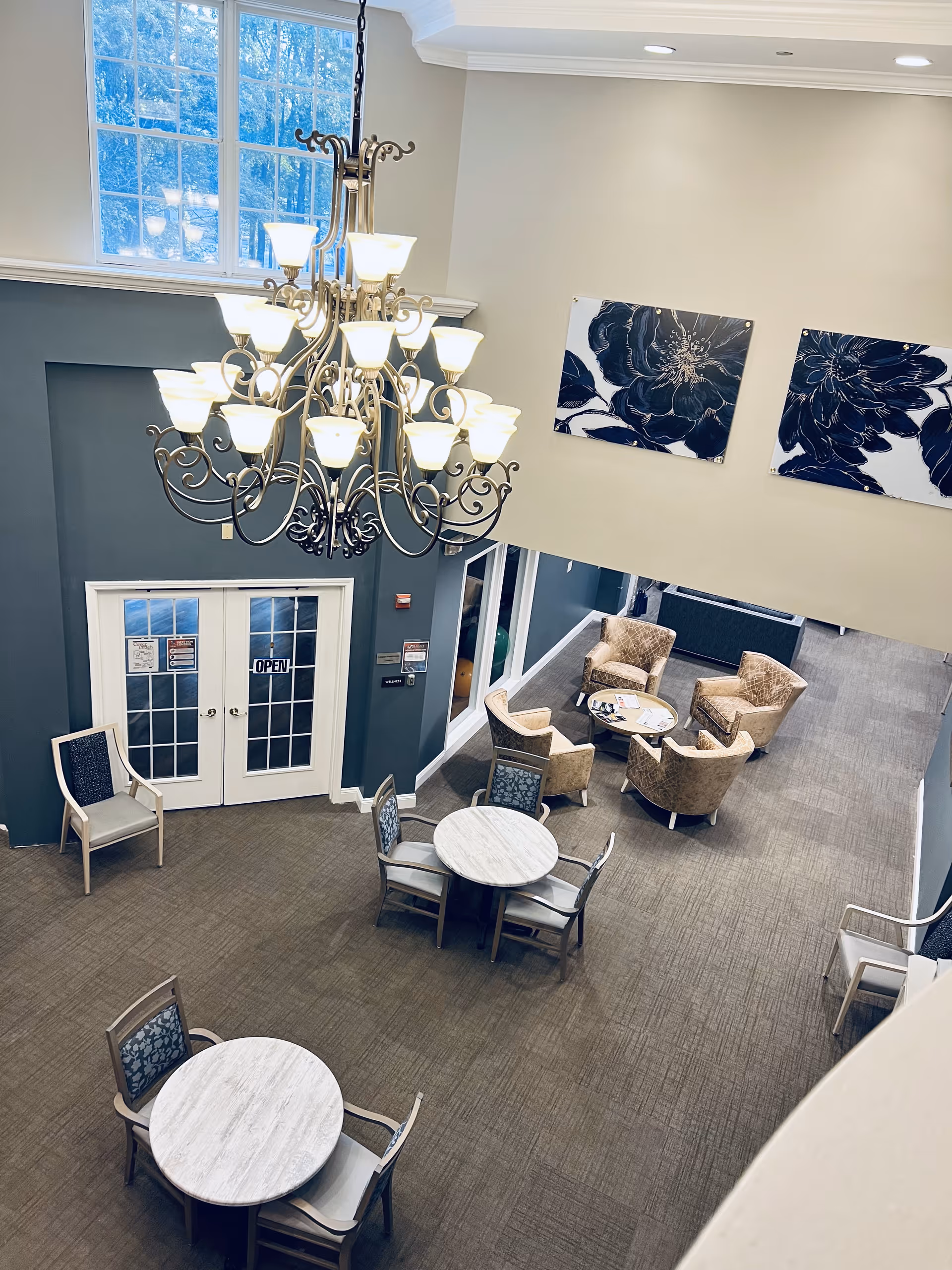 View of a senior living facility common area with round tables and chairs, a seating area with four armchairs around a small round table, a large chandelier hanging from the ceiling, and two large floral paintings on the wall.
