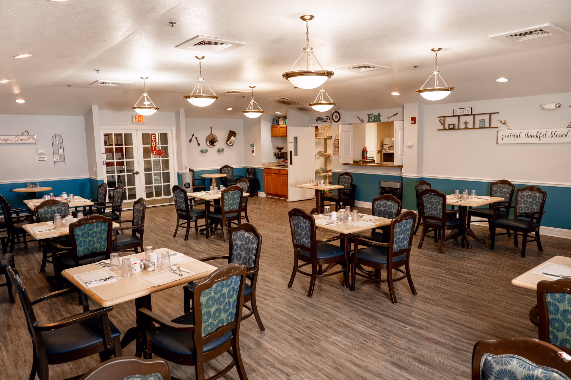 A dining room with multiple tables and chairs arranged neatly. Each table is set with placemats, utensils, cups, and glasses. The room has wood flooring, teal and white walls, and ceiling lights. There is a serving window and kitchen area visible in the background, along with decorative wall signs and a door with glass panels.