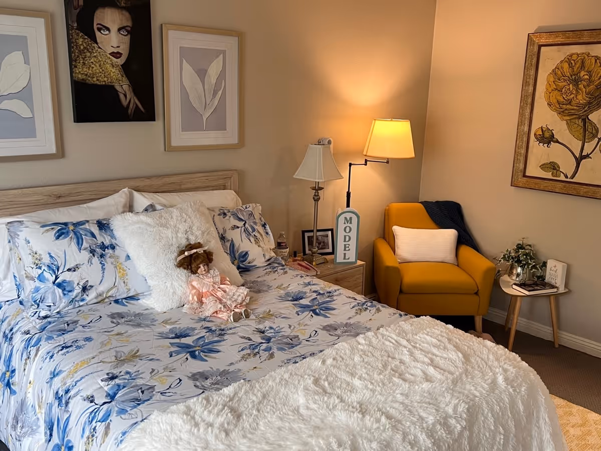 A cozy bedroom with a bed featuring floral blue and white bedding, a fluffy white pillow, and a doll dressed in pink sitting on the bed. There are three framed artworks on the walls, a wooden nightstand with two lamps and a 'MODEL' sign, and a mustard yellow armchair with a white pillow and a dark throw blanket. A small round table next to the armchair holds a plant and a book.