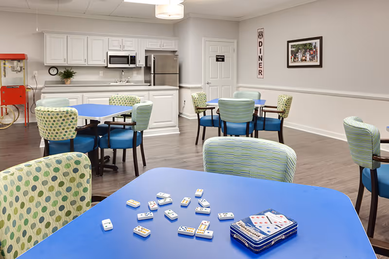 Bright communal dining and activity room with blue tables and patterned chairs, a kitchenette in the back, and dominoes on the foreground table.