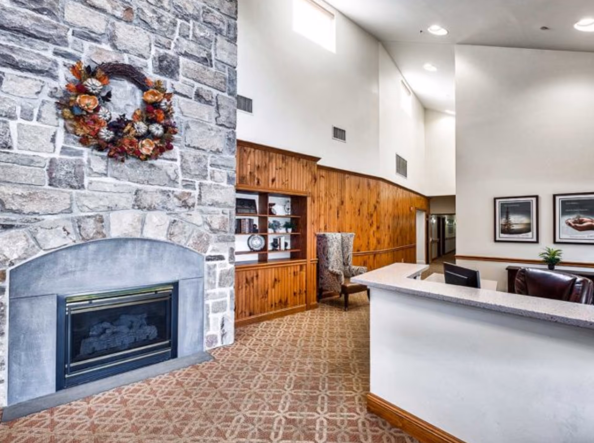 Interior view of a senior living facility lobby featuring a large stone fireplace decorated with a fall-themed wreath, a wooden built-in bookshelf with decorative items, a patterned carpet, a high ceiling with recessed lighting, a reception desk with a computer monitor, and framed artwork on the wall.