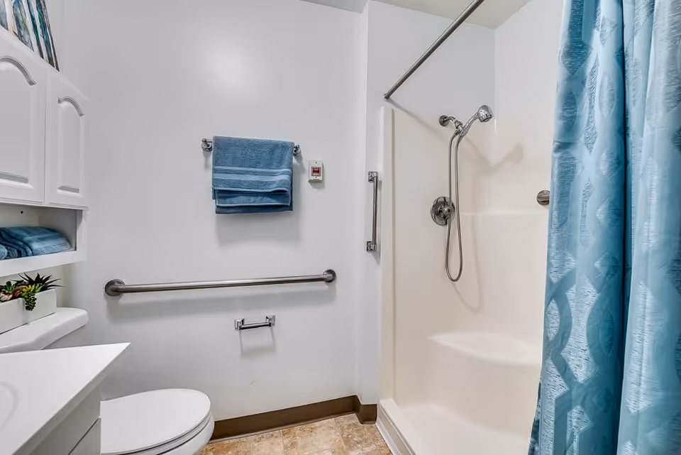 A clean and accessible bathroom with a white toilet, white sink cabinet, and a shower area with a blue patterned shower curtain. There are blue towels hanging on a towel rack and a grab bar installed on the wall next to the shower. The floor has beige tiles and the walls are white.
