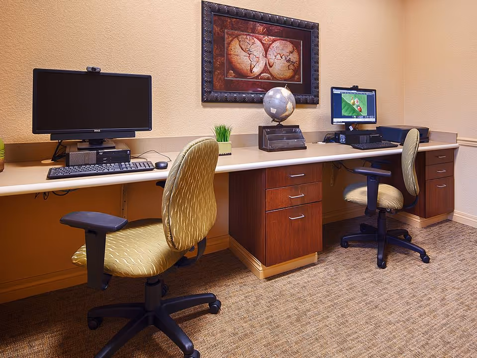 A small office area with two computer workstations, each with a desktop computer, keyboard, and mouse. Two green patterned office chairs are positioned in front of the desks. On the desk between the computers is a decorative globe and a small potted plant. A framed vintage world map hangs on the beige wall above the desk. The floor is carpeted in a neutral tone.