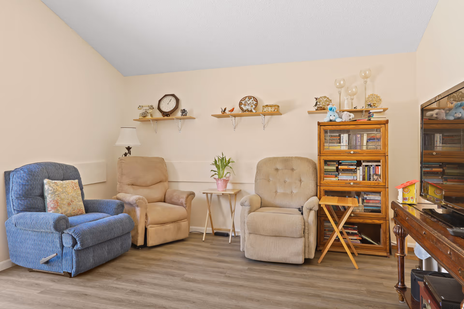 A cozy living room with three recliner chairs in blue, beige, and light brown colors arranged against a beige wall. There are three wooden shelves mounted on the wall displaying decorative items including a clock, figurines, and glass candle holders. A small wooden side table with a potted plant is placed between the chairs. To the right, there is a wooden bookcase filled with books and topped with stuffed animals and more decorative items. A flat-screen TV is partially visible on the right side on a wooden stand.