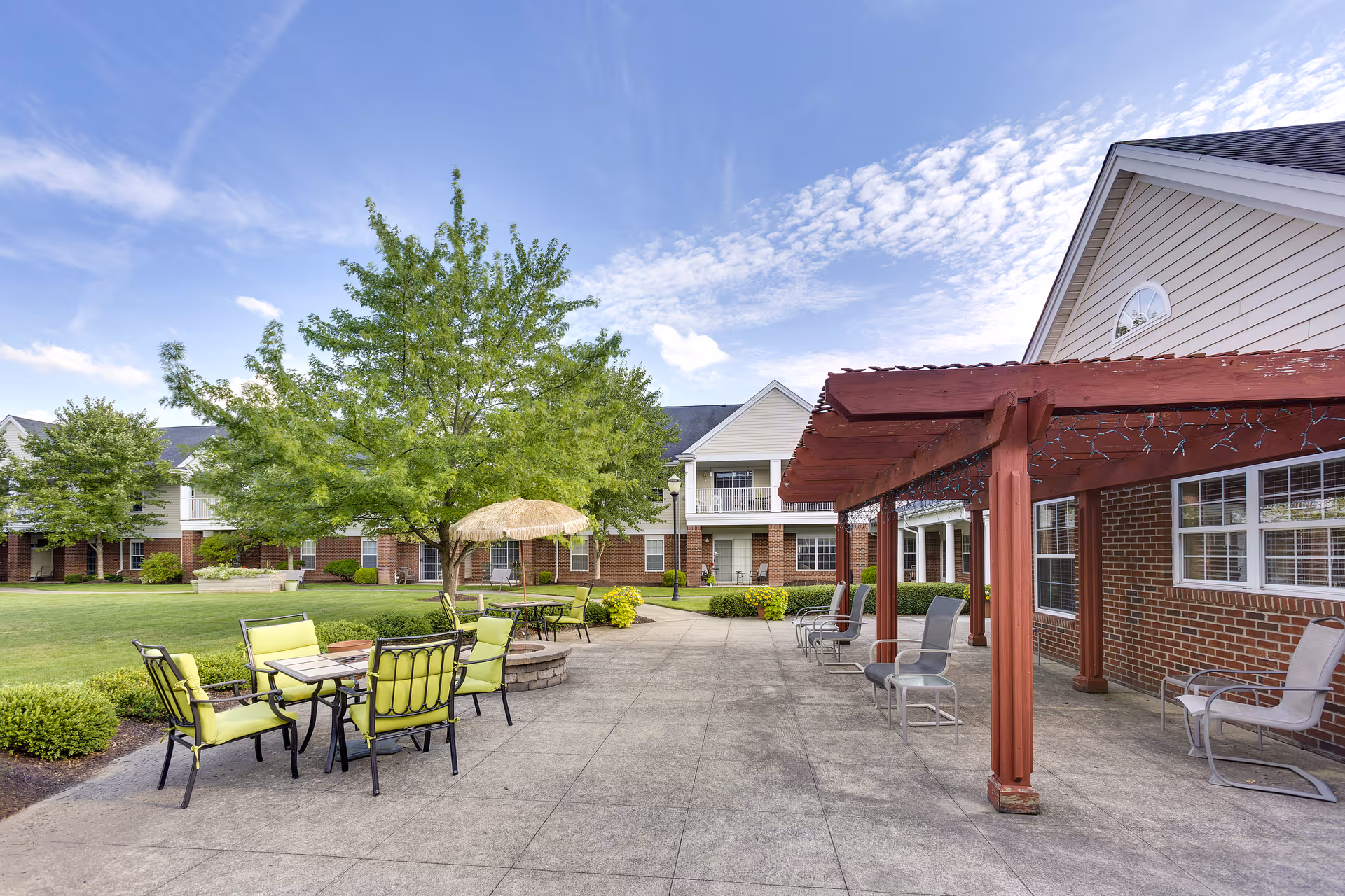 Outdoor patio area at Independence Village of Aurora with green cushioned chairs around tables, a fire pit, a large tree, and a red pergola attached to a brick building under a partly cloudy sky.