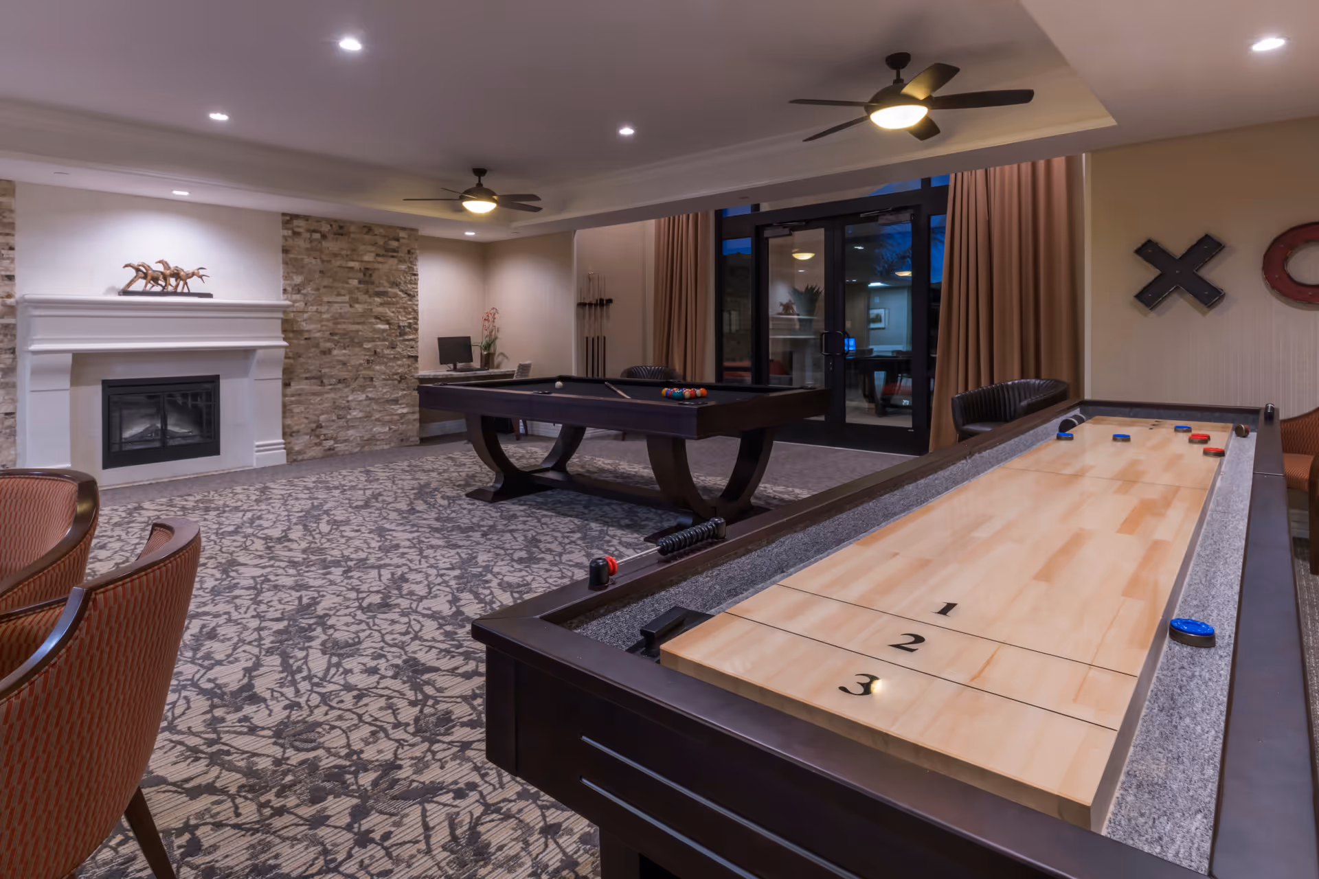 A senior living facility game room featuring a shuffleboard table in the foreground and a pool table in the background. The room has a patterned carpet, a stone accent wall with a fireplace, ceiling fans with lights, and large glass doors with curtains. There are also chairs and wall decorations including large tic-tac-toe X and O symbols.