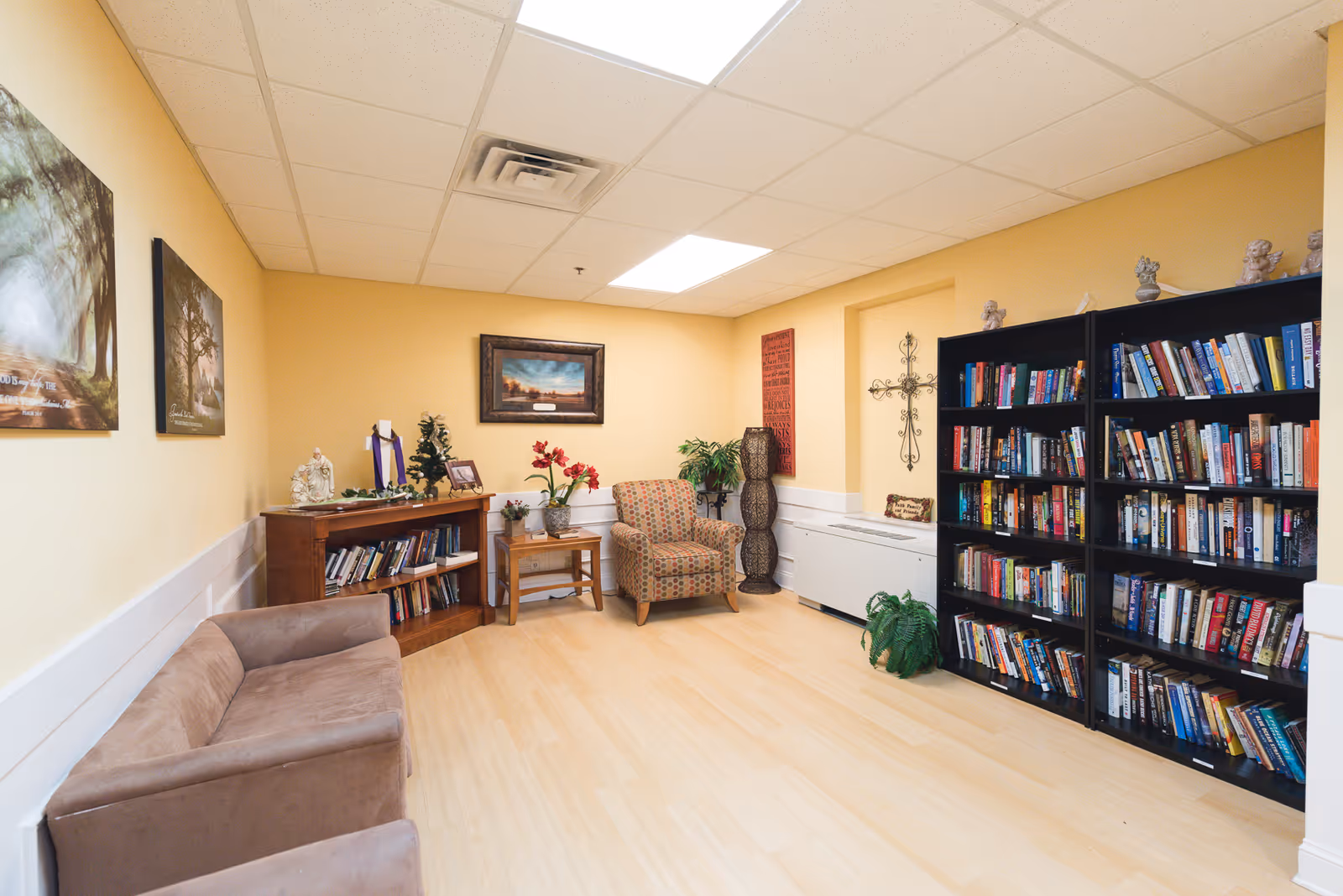 A cozy room with light yellow walls and a light wood floor, featuring a brown couch on the left, a patterned armchair in the corner, a wooden bookshelf filled with books, a black bookshelf filled with books on the right, decorative plants, framed artwork on the walls, and a decorative cross.