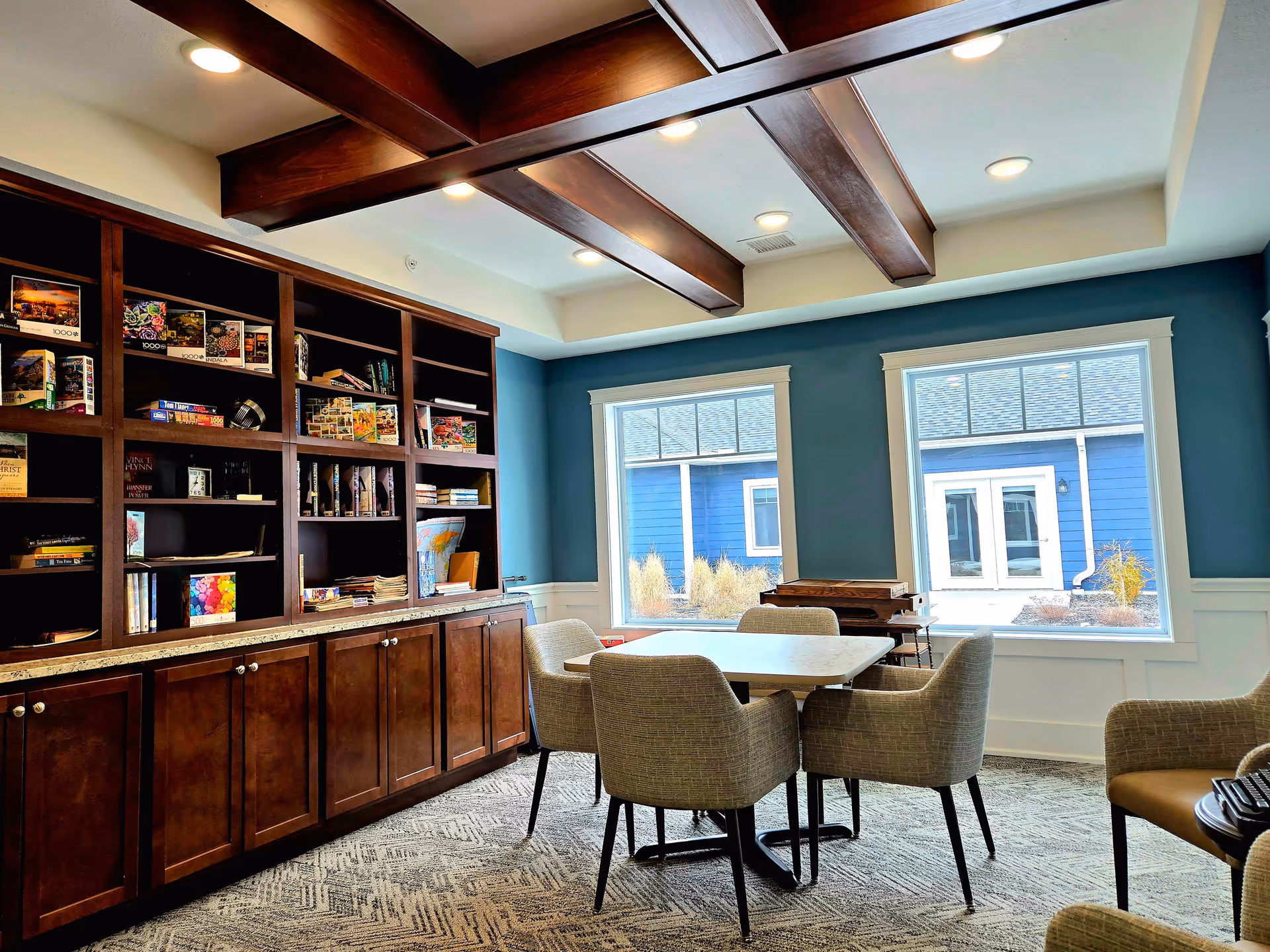 Bright common room with built-in bookshelves, a table surrounded by upholstered chairs, and large windows overlooking a blue building.
