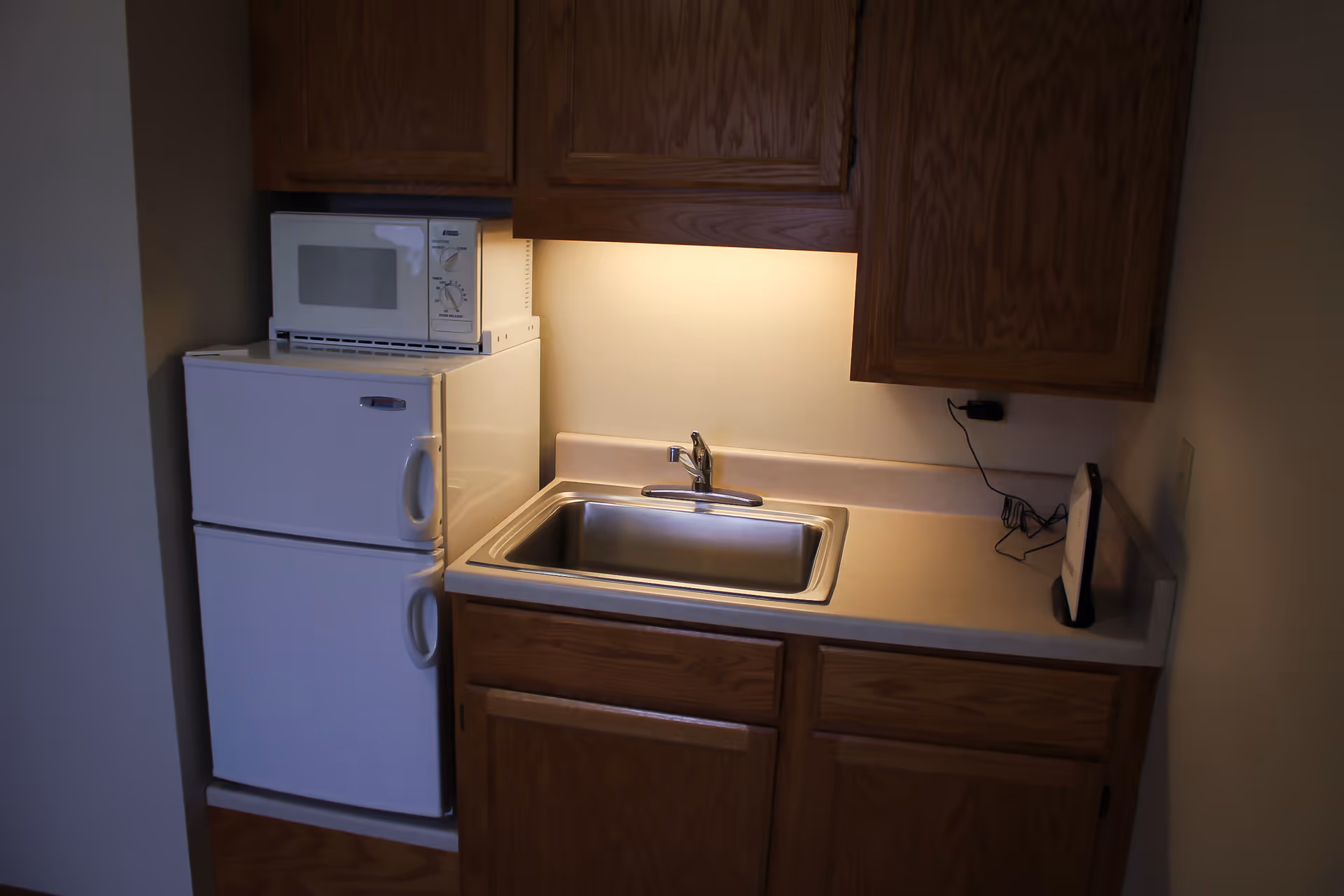 Small kitchenette area with wooden cabinets, a white mini refrigerator with a microwave on top, a stainless steel sink, and a countertop with a small electronic device plugged into the wall.