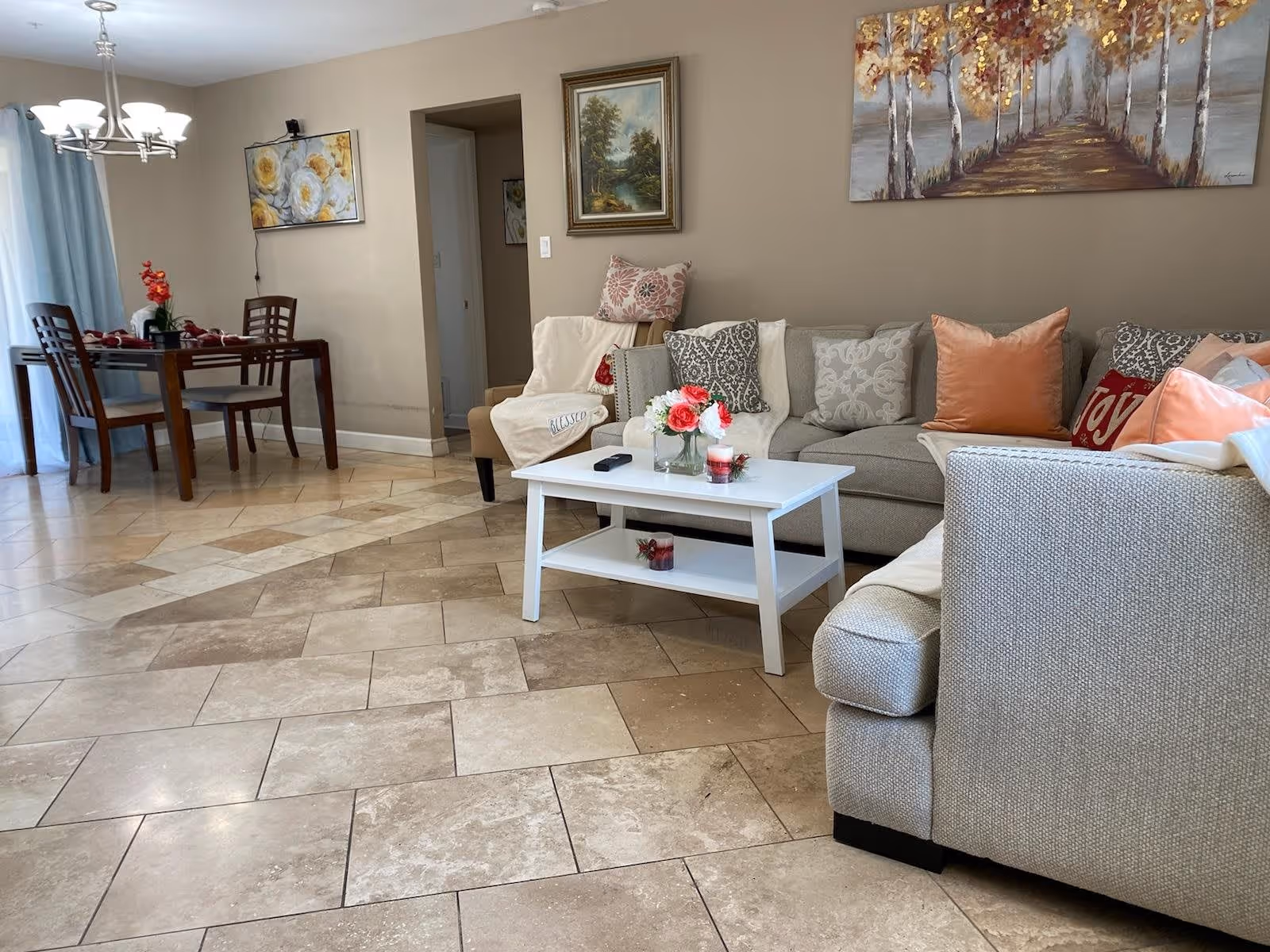 A cozy living room and dining area with beige tiled floors. The living room features a light gray sectional sofa adorned with various decorative pillows and a white coffee table with flowers and candles. The dining area has a wooden table with four chairs and a chandelier overhead. The walls are decorated with framed paintings and the room is softly lit by natural light coming through a window with light blue curtains.